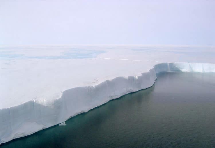 Aerial view of an ice shelf meeting the ocean, showcasing a sharp cliff of ice above dark blue water under a bright sky.